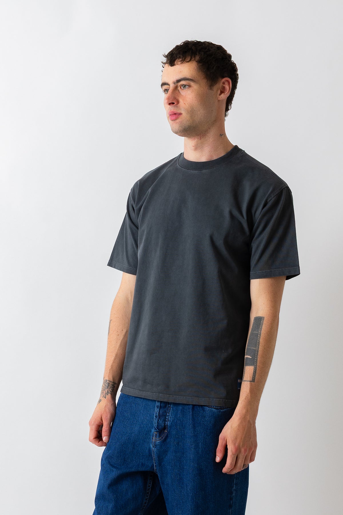 A man wearing a dark charcoal gray crewneck t-shirt and blue jeans, standing against a light gray studio background.