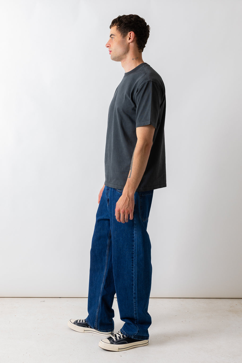 A man in profile wearing a dark charcoal gray t-shirt, wide-leg blue jeans, and black canvas sneakers, standing against a plain white studio background.