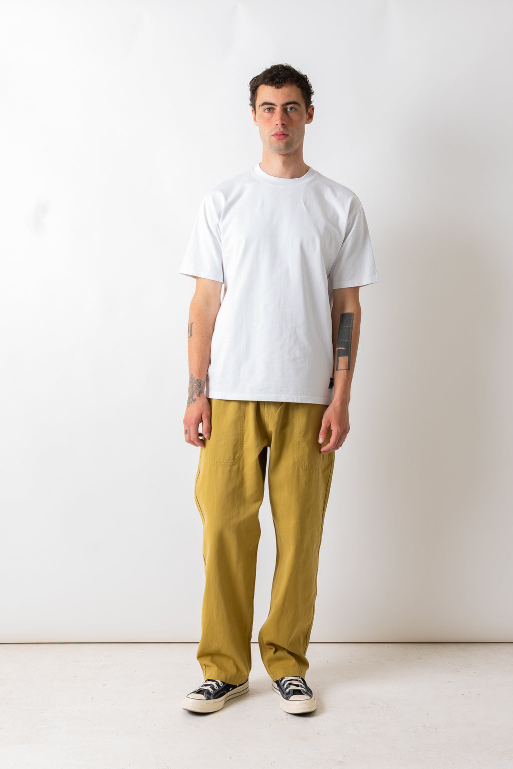 A man stands against a plain off-white studio wall wearing a plain white short-sleeve t-shirt, loose mustard yellow pants, and black-and-white sneakers.