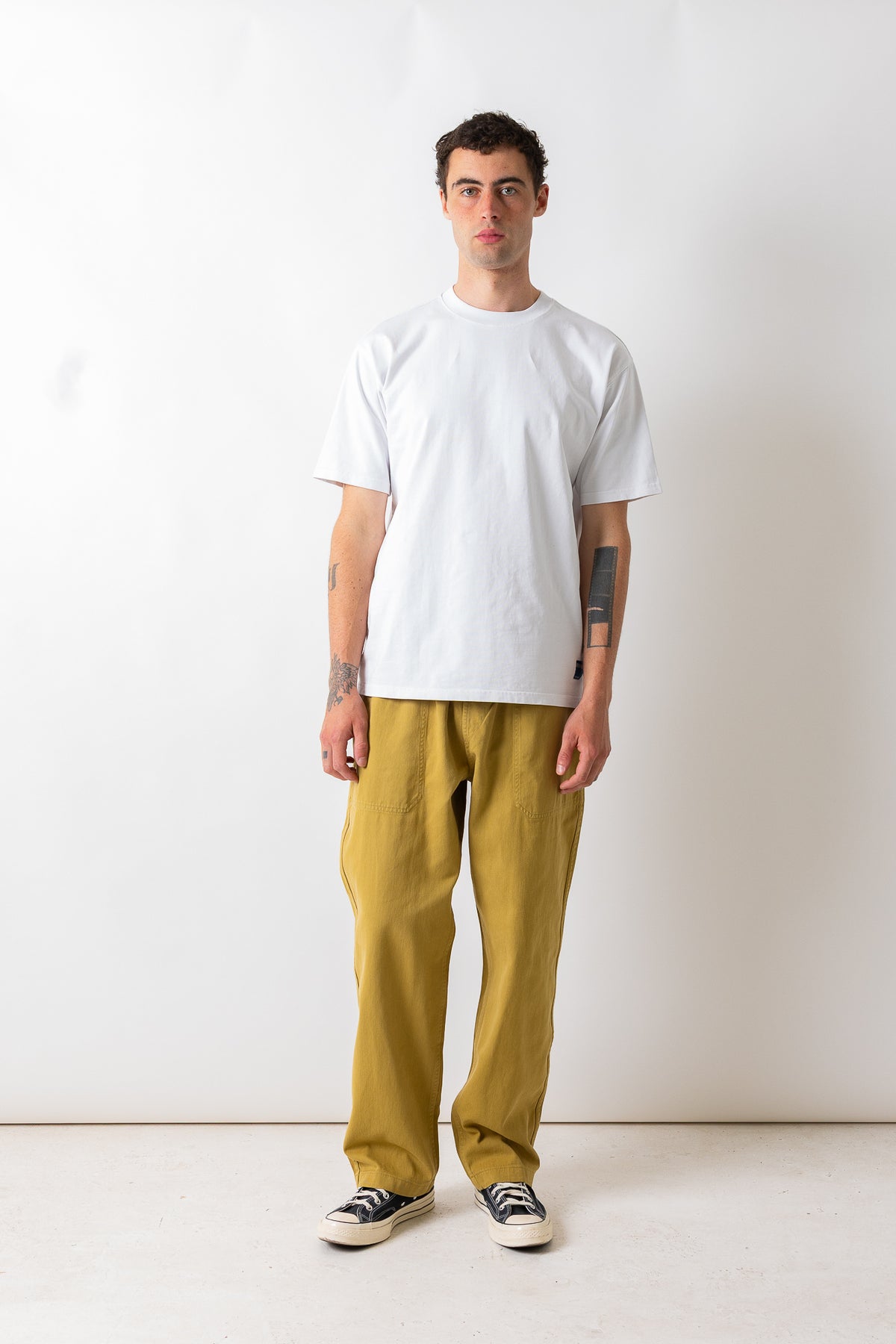 A man stands against a plain off-white studio wall wearing a plain white short-sleeve t-shirt, loose mustard yellow pants, and black-and-white sneakers.