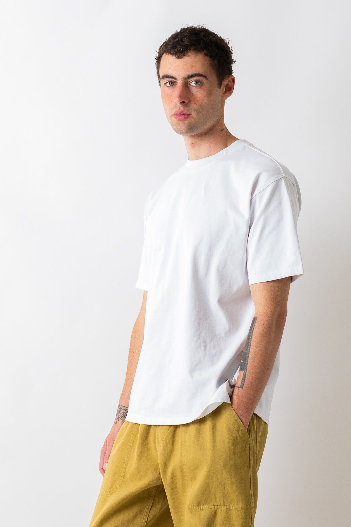 A man wearing a plain white short-sleeve crewneck t-shirt and mustard yellow pants, standing against a light gray studio background.