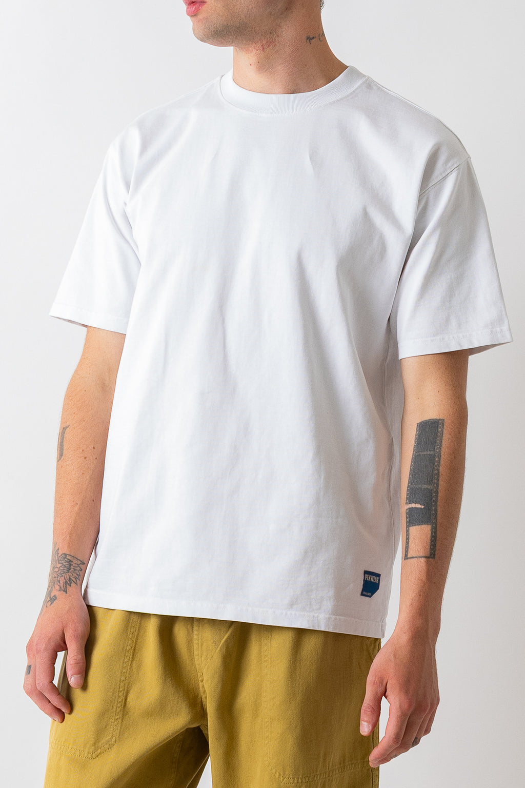 A man wearing a plain white short-sleeve crewneck t-shirt and mustard yellow pants, shown from the chin down against a light gray studio background.