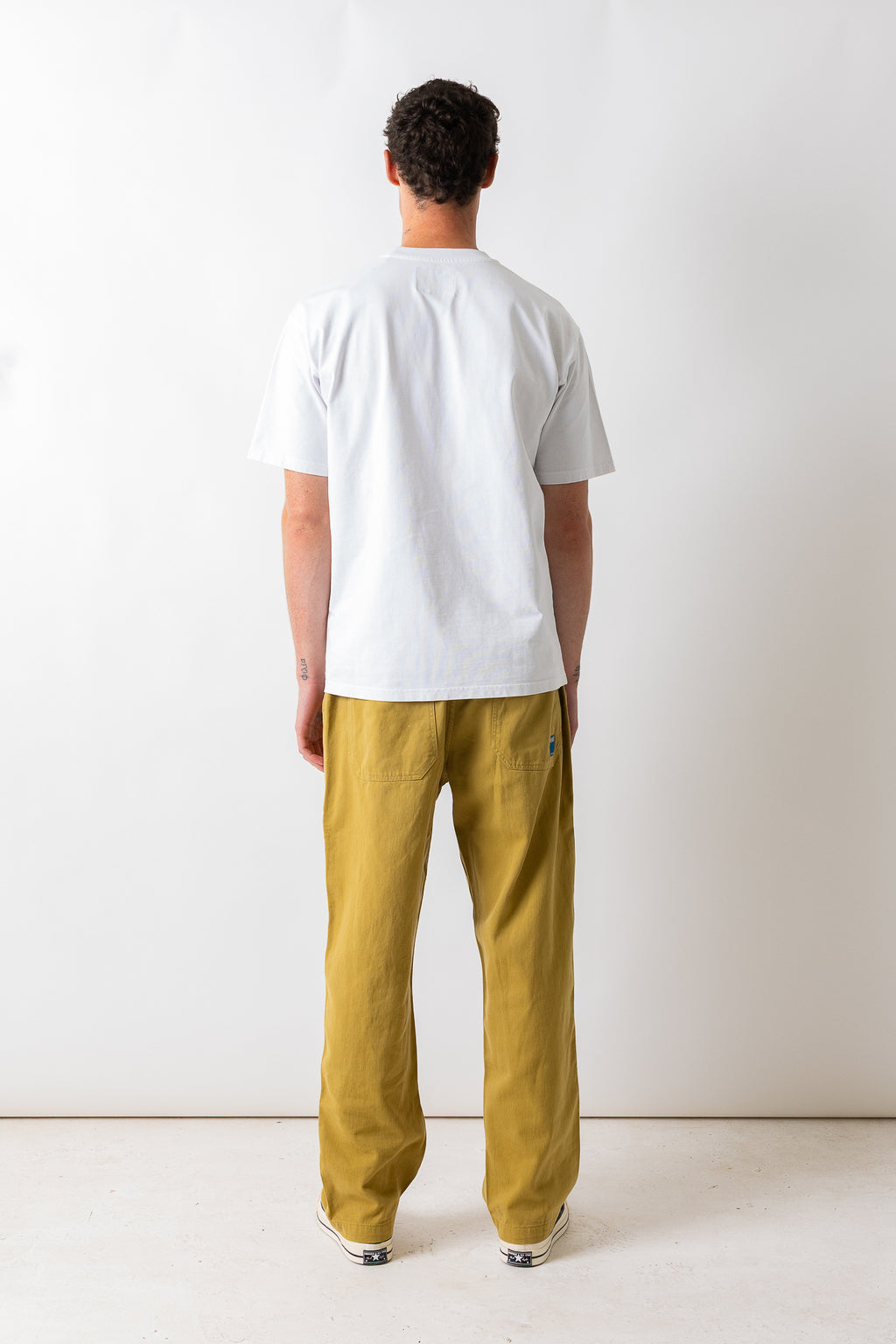 A man viewed from behind wearing a white short-sleeve t-shirt and mustard yellow trousers, standing against a plain white studio background.