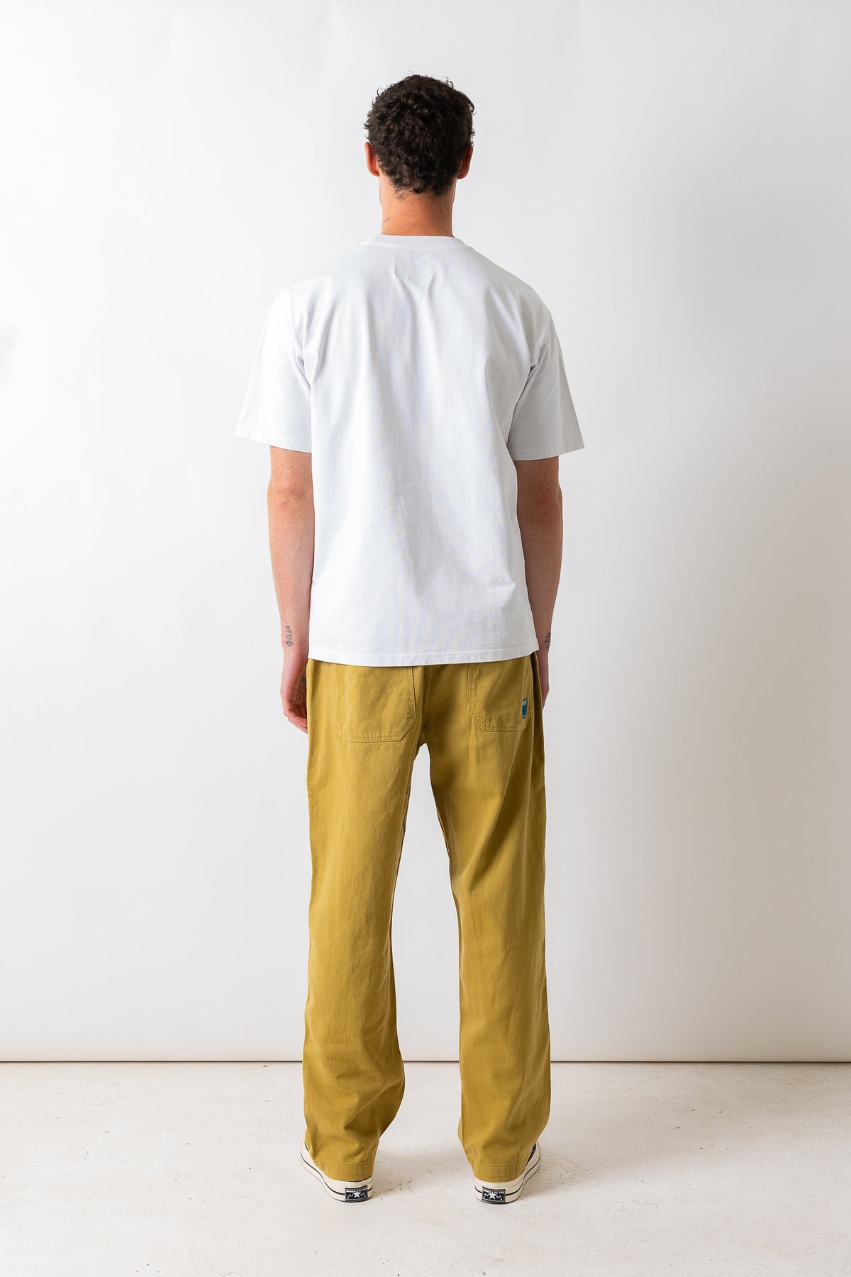 A man viewed from behind wearing a white short-sleeve t-shirt and mustard yellow trousers, standing against a plain white studio background.