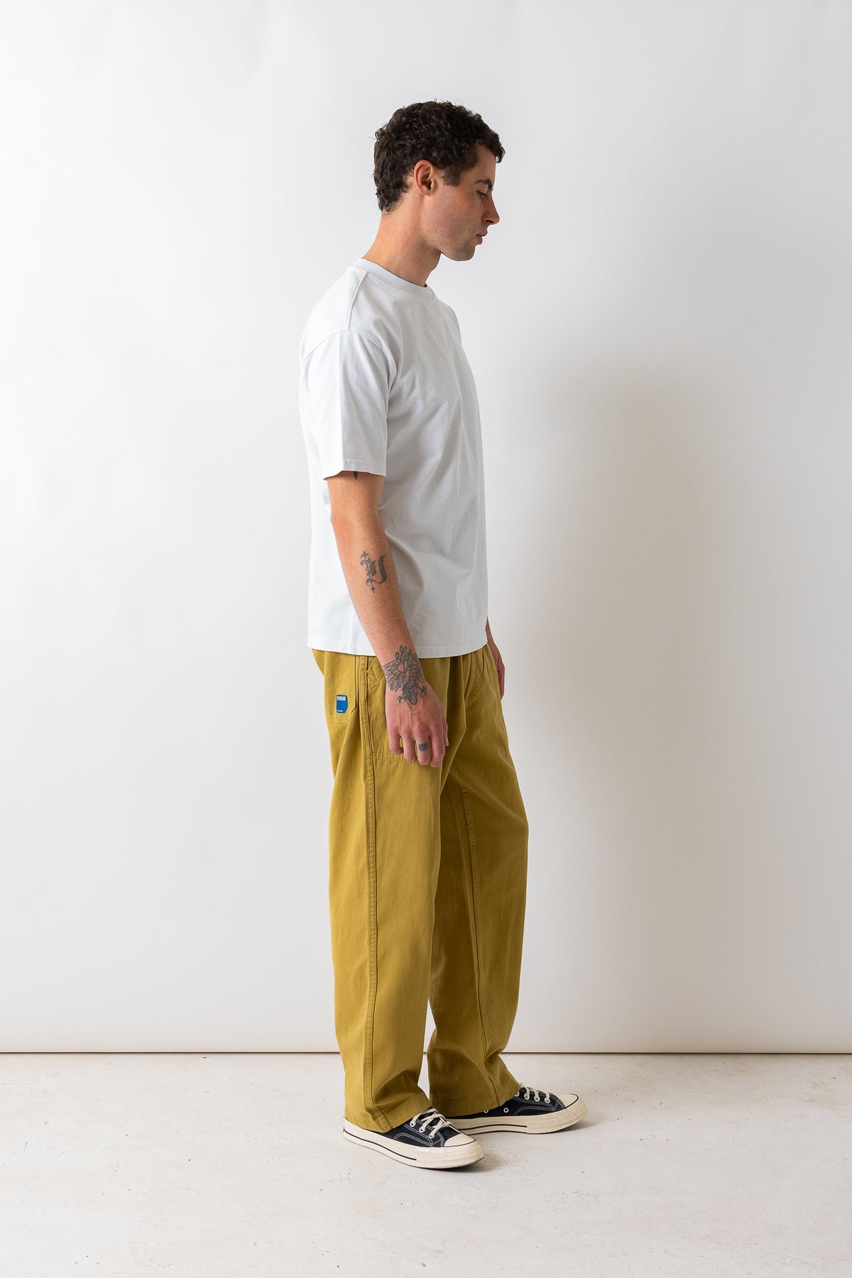 A man in profile wearing a white t-shirt, mustard yellow wide-leg pants, and black canvas sneakers, standing against a plain off-white studio wall.